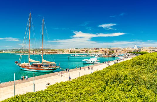 Beautiful view of a promenade in Syracuse with walking people. Cityscape of Ortygia Island with harbor, embankment, park and ships