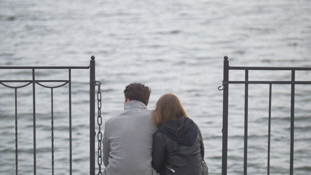Young couple sitting on the pier above river admiring nature in cloudy cold day, close up