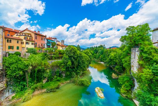 Old Italian city of Cividale del Friuli. Medieval town in Northern Italy. Cityscape with colored residential houses, river, cliffs with green trees and blue sky with clouds.