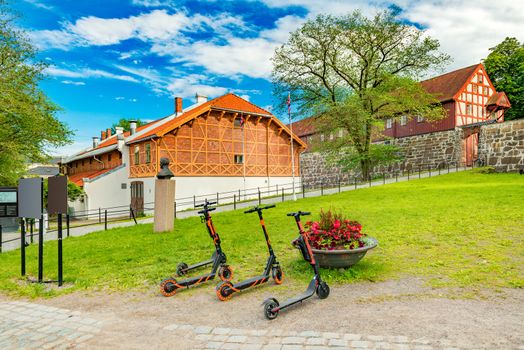 Three electric scooters stand on the grass in front of historical buildings in the old town of Oslo, Norway