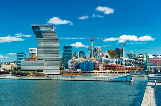 Cityscape of Oslo with modern architecture, buildings under construction, tower cranes, harbor and bridge with walking people, Norway