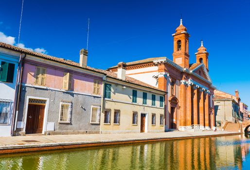 View of the street with canal and colored houses in a small Italian town of Comacchio (Little Venice). San Camillo Hospital (museum of the Roman Ship) against the clear blue sky, Emilia-Romagna, Italy