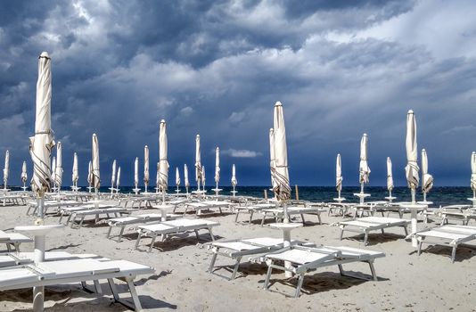 Dark and stormy sky, with gray clouds on the blue sea and a little beach with deck chairs and closed white umbrellas in the foreground