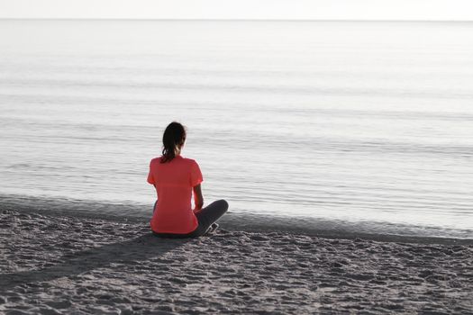 Young woman sitting on the shoreline of the sea at dawn in athletic outfit, looks towards the sea illuminated by the low sun and meditates