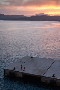 Sunrise on the Sardinian sea coast with intense orange color seen from the sea on the ferry that is about to dock and two workers of the port on the pier