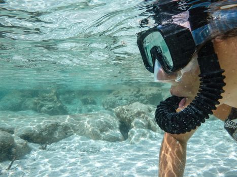 Close up of the face of a young woman in bikini with mask and snorkel shooting underwater in the crystal clear sea with white sand and rocks on the bottom