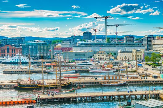 Oslo - June 2019, Norway: Aerial view of the harbor of Oslo with modern and historical ships. Cityscape with modern port, architecture, tower cranes, piers with walking people, hills and the blue sky