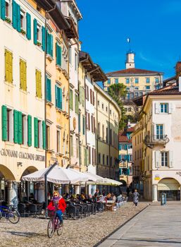 Udine - February 2017, Northern Italy: Woman in red jacket is cycling in the center of the city. The street of Udine with colored houses in traditional architectural style. People are sitting in cafe