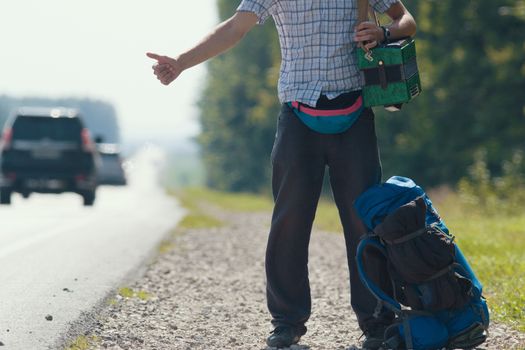 A man in a plaid shirt, dark pants, with an accordion on his shoulders standing by the road and honking, backpack lying at his feet, close-up