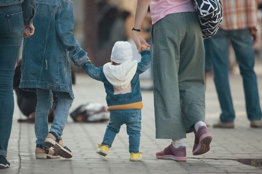 Cute baby walking with his mother and sister on the street, holding hands mom and sister, close up