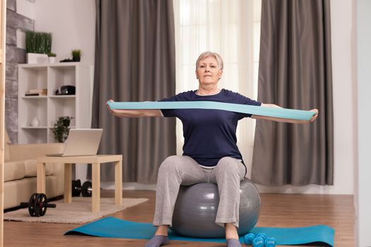 Ambitious woman doing sports at home with resistance band. Old person pensioner online internet exercise training at home sport activity with dumbbell, resistance band, swiss ball at elderly retirement age