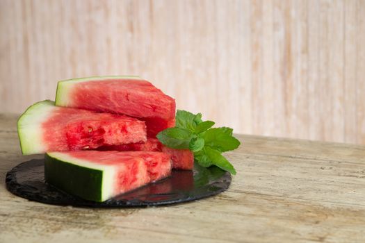 Triangular slices of watermelon overlaid on a black plate of wet slate with sprig of fresh green mint in selective focus for copy space