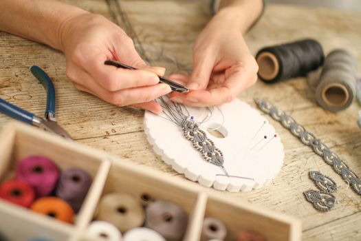 Close up of female hands making a macramé bracelet with kumihimo on a wooden table with tools, spools of thread, natural stones and colored beads