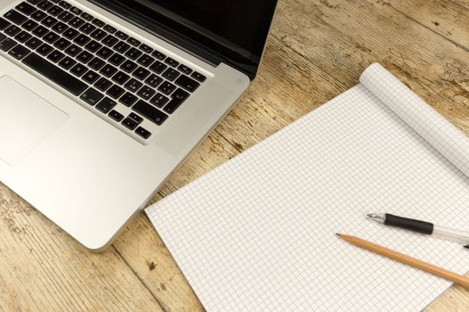 Contrast between old and new office work: shot from the top of an aluminum laptop on a wooden desk with an open checkered notebook, a pencil and a pen