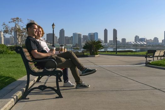 Young couple is sitting on a bench in the city, the sun is shining, they are talking and laughing, watching the distance and dreaming. In the background is the downtown San Diego with skyscrapers.