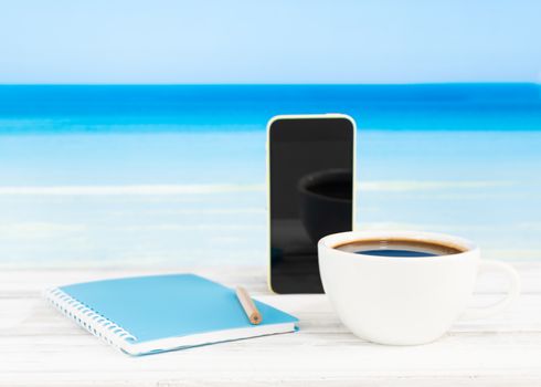Coffee cup,mobile phone and blue diary on white wooden table with summer beach background