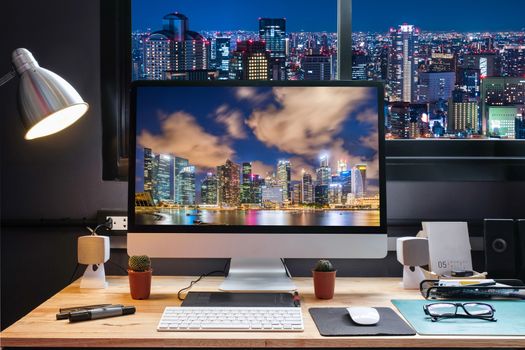 Graphic designer's workspace equipped with a pen tablet, a computer and cityscape screen for text with beautiful night view city from the window 