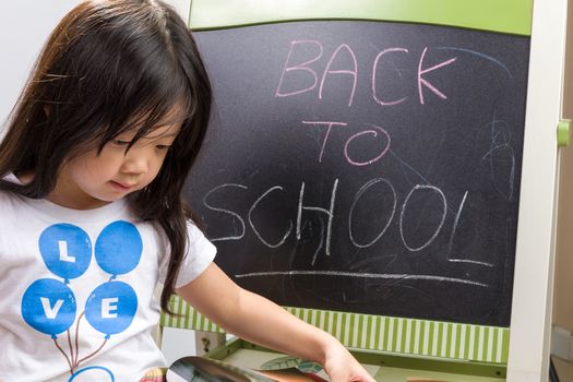 Child holding book in her hands beside blackboard to illustrate education concept.