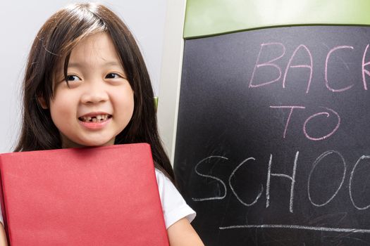 Child holding book in her hands beside blackboard to illustrate education concept.