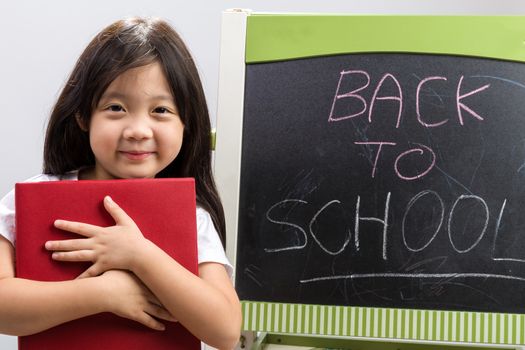 Child holding book in her hands beside blackboard to illustrate education concept.