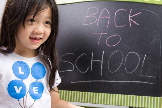 Child holding book in her hands beside blackboard to illustrate education concept.