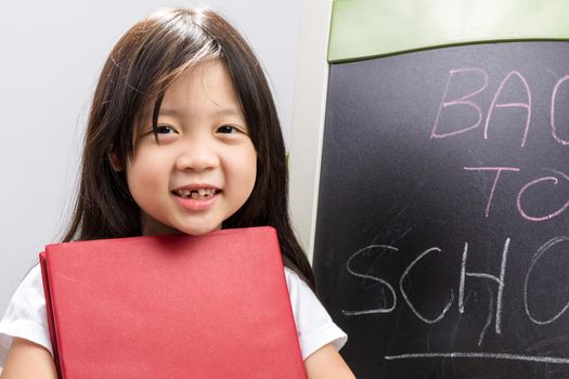 Child holding book in her hands beside blackboard to illustrate education concept.