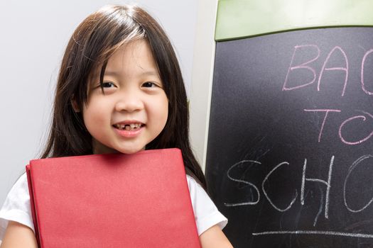 Child holding book in her hands beside blackboard to illustrate education concept.