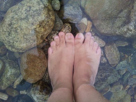 Top view of transparent water surface with human feet visible in it. Copy space for background and texture. Transparent water texture with view of stones under water.