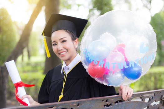 Portrait of happy young female graduates in academic dress and square academic cap holding word quotes of CONGRATS GRAD on balloon after convocation ceremony.