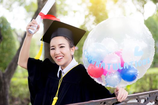 Portrait of happy young female graduates in academic dress and square academic cap holding word quotes of CONGRATS GRAD on balloon after convocation ceremony.