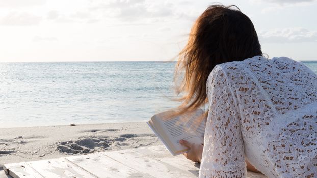 The young woman from behind lying by the sea looks at the horizon at dawn in the wind, dressed in a white lace dress and white underwear and long hair