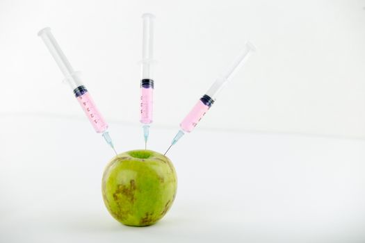 Concept: human GMO manipulation of nature and relative poisoned fruits. Close-up of an apple contaminated with three syringes threaded in and white background