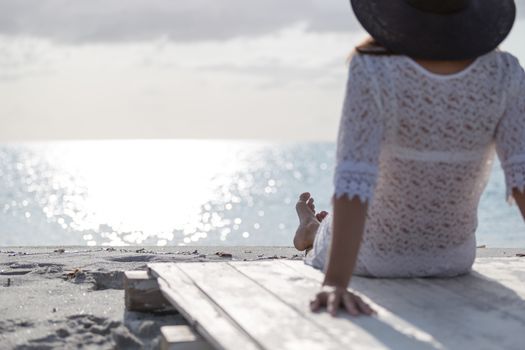 Young woman with long hair from behind sitting by the sea looks at the horizon at dawn in the wind, dressed in a white lace dress, white underwear and large black hat