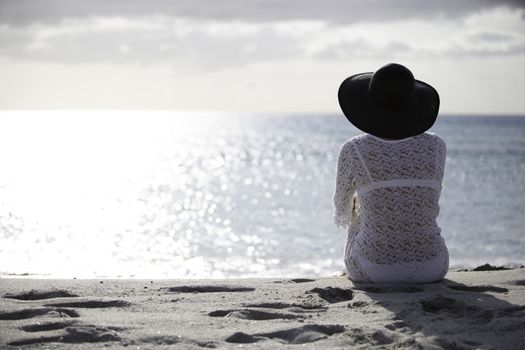 Young woman with long hair from behind sitting by the sea looks at the horizon at dawn in the wind, dressed in a white lace dress, white underwear and large black hat