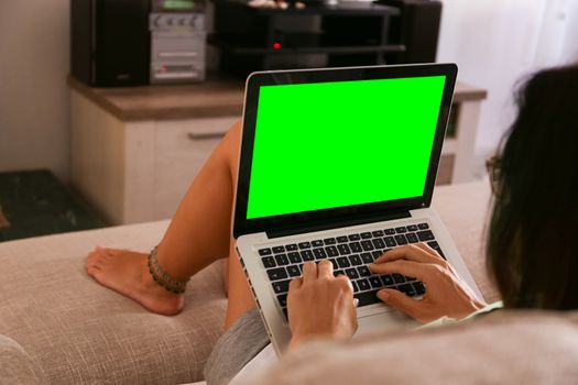 A young woman works on her laptop with the green screen at home sitting on the sofa in her living room