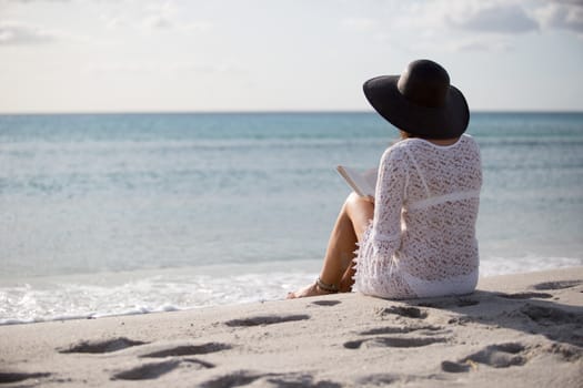 Young woman with long hair from behind sitting by the sea reads a book at dawn in the wind, dressed in a white lace dress, white underwear and large black hat