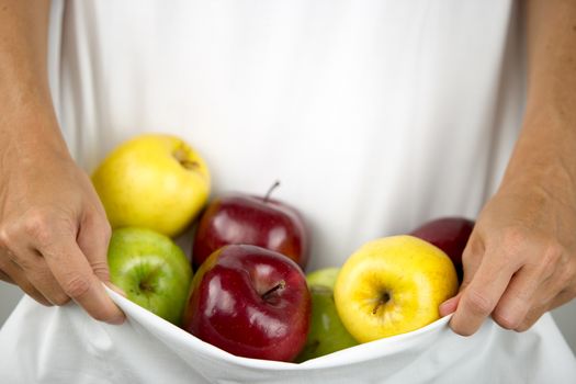 A Caucasian woman holds some apples of various types and colors in her white dress clasped in her hands