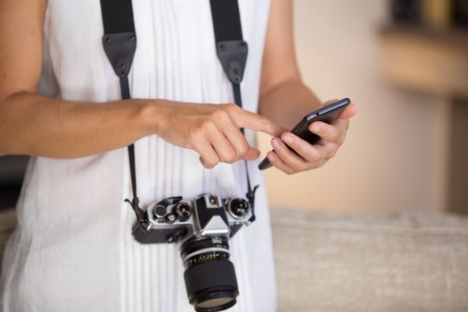 Contrast between old and modern times: a young woman with a vintage camera around her neck fiddles with her smartphone