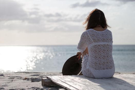 Young woman from behind sitting by the sea looks at the horizon at dawn in the wind, dressed in a white lace dress and white underwear and long hair