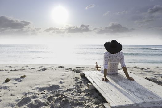 Young woman with long hair from behind sitting by the sea looks at the horizon at dawn in the wind, dressed in a white lace dress, white underwear and large black hat