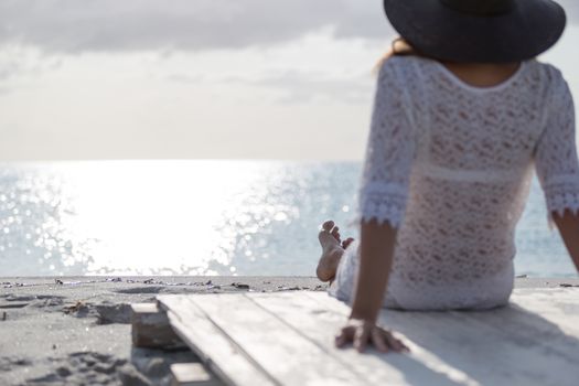 Young woman with long hair from behind sitting by the sea looks at the horizon at dawn in the wind, dressed in a white lace dress, white underwear and large black hat