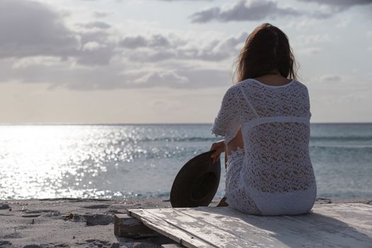 Young woman from behind sitting by the sea looks at the horizon at dawn in the wind, dressed in a white lace dress and white underwear and long hair