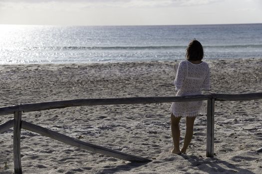 Young woman from behind reads a book on the beach looking at the horizon at dawn in the wind, dressed in a white lace dress, white underwear and long hair