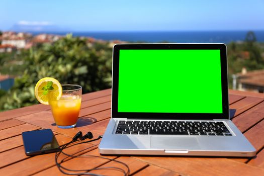 Open laptop with green screen on wooden table with the smartphone and the connected headphones and the blue sea panorama in the background in selective focus