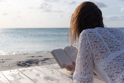 The young woman from behind lying by the sea looks at the horizon at dawn in the wind, dressed in a white lace dress and white underwear and long hair