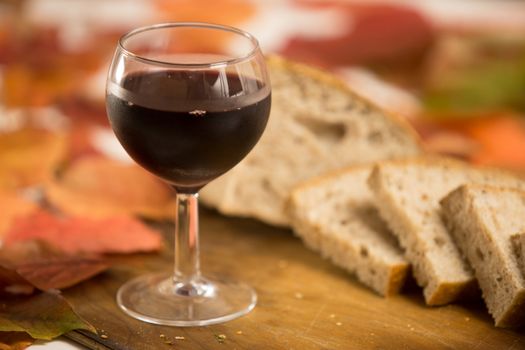 Autumn flavors and colors: a glass of red wine with slices of wholemeal bread in the background on an old wooden cutting board with red and orange wisteria leaves around
