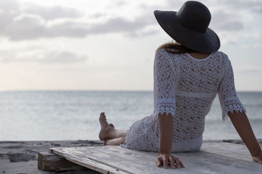 Young woman with long hair from behind sitting by the sea looks at the horizon at dawn in the wind, dressed in a white lace dress, white underwear and large black hat