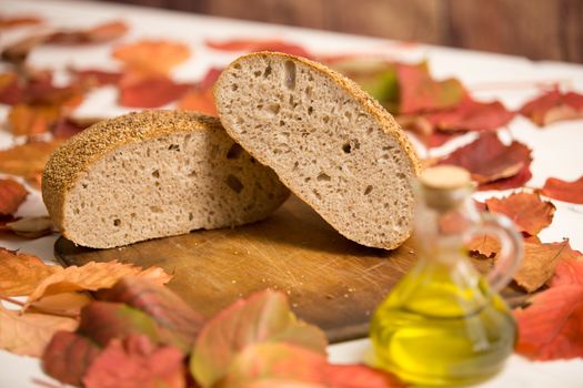 Autumnal rustic food: a whole loaf cut in half on an old wooden cutting board, ampoule of extra virgin olive oil and red and orange wisteria leaves around