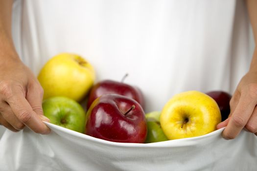 A Caucasian woman holds some apples of various types and colors in her white dress clasped in her hands