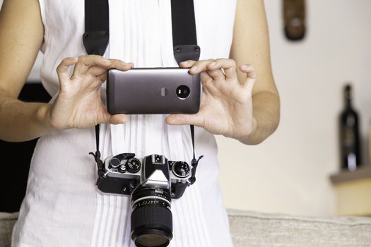 Contrast between old and modern times: a young woman with a vintage camera around her neck fiddles with her smartphone